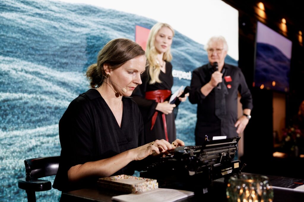 Nicola Gold an Schreibmaschine auf Bühne, im Hintergrund Moderatorin Larissa Robitschko und Viereck-Gründer Ewald Viereck